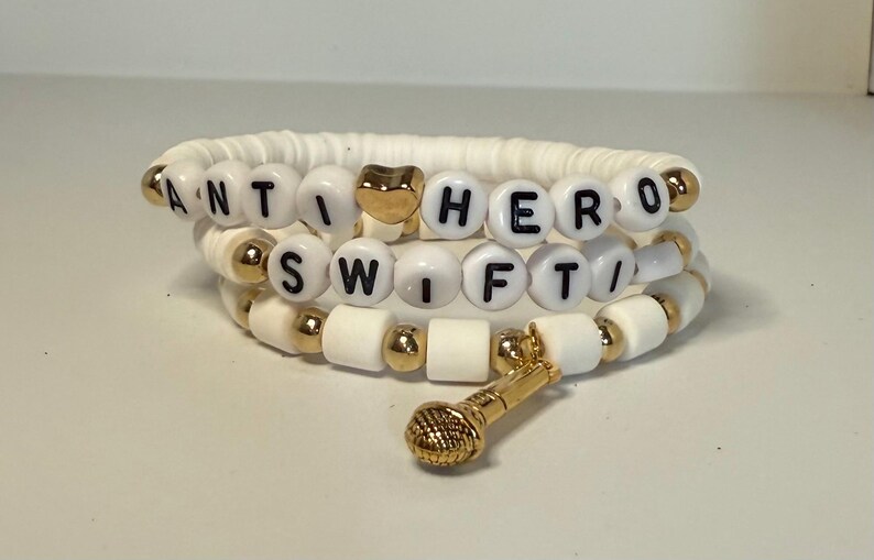 May include: Three-strand bracelet with white beads and gold accents. The bracelet spells out "ANTI HERO SWIFT" with black letter beads. A gold heart bead and a gold microphone charm are also included.