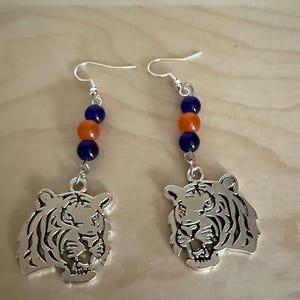 May include: A pair of silver-colored tiger head earrings. Each earring features a tiger head pendant, with a series of blue and orange beads. The earrings have silver-colored ear wires.