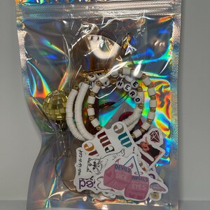 May include: A clear, holographic zip bag containing various accessories. Inside are white and gold beaded bracelets spelling "HERO", stickers with text and graphics, and a gold disco ball charm. The bag is sealed.