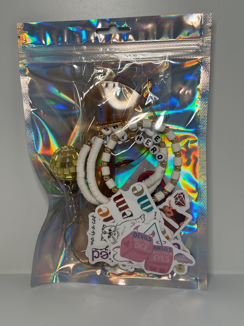May include: A clear, holographic zip bag containing various accessories. Inside are white and gold beaded bracelets spelling "HERO", stickers with text and graphics, and a gold disco ball charm. The bag is sealed.