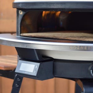 May include: A black and cream-colored outdoor pizza oven with a stainless steel trim. The oven features a digital display and a temperature control knob. The oven is designed for outdoor cooking and has a wooden shelf.