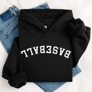 Puede incluir: Sudadera negra con la palabra "BASEBALL" en letras blancas arqueadas. La sudadera tiene un cordón y una capucha. Las mangas están dobladas. La sudadera está colocada sobre unos vaqueros azules.