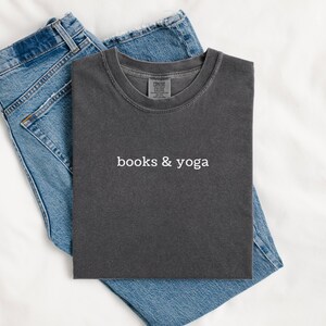 May include: A charcoal gray crew neck t-shirt featuring the white text "books & yoga" printed on the chest, layered over a pair of blue denim jeans. The shirt has a faded, vintage-style wash and a standard short-sleeve construction.