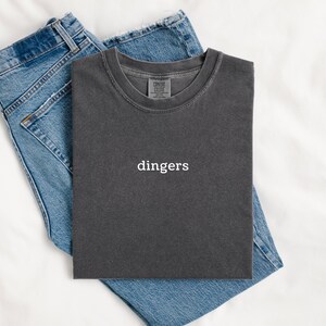 May include: A charcoal gray t-shirt with the word "dingers" in white, laid on top of a pair of blue denim jeans. The t-shirt has a crew neck and a vintage, washed look. The jeans are folded neatly.