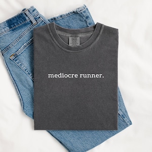 May include: A charcoal grey crew neck t-shirt with the text "mediocre runner." printed in white across the chest, paired with a pair of blue denim jeans. The t-shirt features a ribbed collar and a relaxed fit, showcasing a simple graphic design.