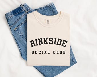 May include: A cream-colored crew neck t-shirt featuring the black text RINKSIDE SOCIAL CLUB in a collegiate-style font, layered over a pair of blue denim jeans. The shirt has a standard short-sleeve design and a simple round neckline.