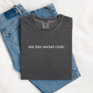 Könnte beinhalten: Ein anthrazitfarbenes T-Shirt mit Rundhalsausschnitt und dem weißen Schriftzug "sin bin social club." auf der Brust, kombiniert mit einer blauen Jeans. Das T-Shirt hat eine Vintage-Waschung und einen klassischen Kurzarm-Schnitt.
