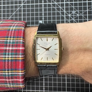 May include: A gold-coloured rectangular quartz watch with a black leather strap. The watch face is light-coloured with black hands and markers. The watch reads "SUNLORD objet QUARTZ". The watch is on a wrist next to a red and black plaid sleeve.