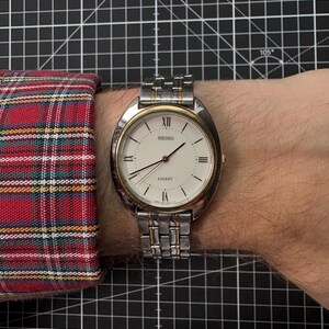 May include: A Seiko Lucent wristwatch with a silver and gold-tone metal band and a white face. The watch has Roman numeral hour markers and a date display. The watch is on a wrist with a plaid shirt sleeve.
