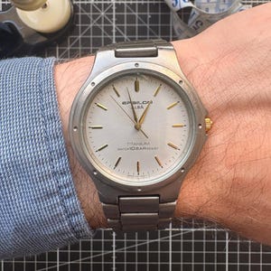 May include: A silver Epsilon Alba titanium watch with a light-coloured face and gold accents. The watch has a metal strap and is water-resistant. The watch is on a wrist, with a blue shirt sleeve visible.