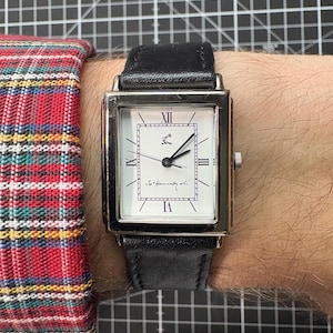 May include: A rectangular wristwatch with a silver case and a black leather strap. The white watch face features black Roman numerals and hands. The watch is on a wrist, with a red and black plaid shirt sleeve visible.