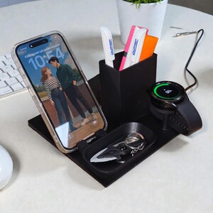 Puede incluir: Un organizador de escritorio negro con soporte para teléfono, reloj y bolígrafos. El teléfono muestra una pareja y el reloj muestra el 50% de batería. El organizador también tiene una pequeña bandeja con llaves.