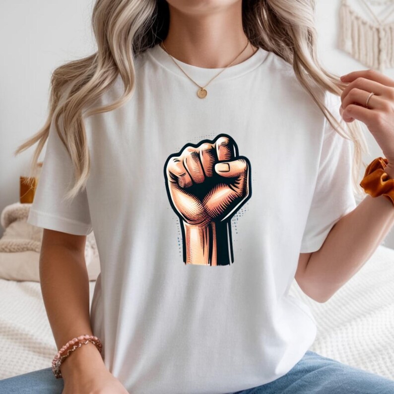 May include: White t-shirt with a graphic of a clenched fist. The fist is illustrated in tan, brown, and black, with detailed shading. The design is centered on the shirt, offering a bold statement.