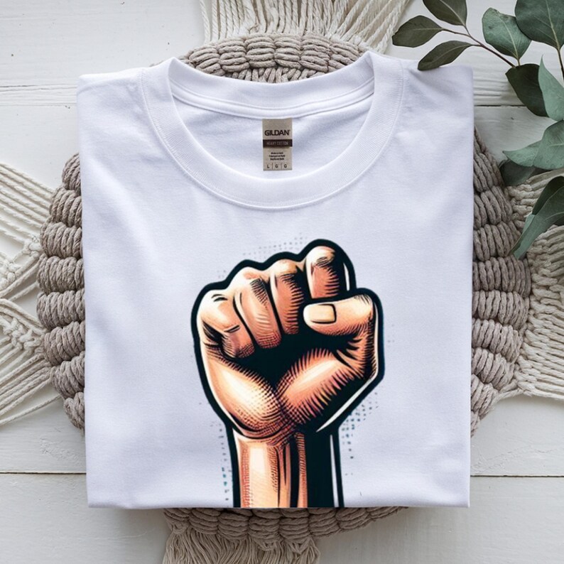May include: White t-shirt with a graphic of a clenched fist in tan, brown, and black. The design is centered on the shirt, with the brand label visible at the top. The shirt is folded on a textured surface.