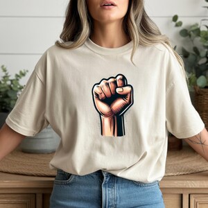 May include: Beige t-shirt with a graphic of a clenched fist. The fist is in shades of orange and brown, with black outlines. The shirt is a neutral colour and has a relaxed fit.
