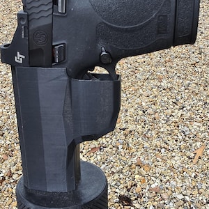 May include: Black handgun in a black holster. The handgun has a red dot sight and is held in a 3D-printed holster with a textured base. The background is a gravel surface.