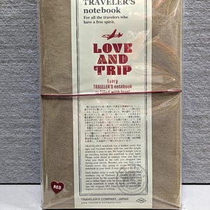 Może przedstawiać: Brązowy notatnik Traveler's, zapakowany w przezroczysty plastik, z czerwoną gumką. Okładka zawiera tekst "TRAVELER'S notebook" i "LOVE AND TRIP". Widoczna jest czerwona naklejka w kształcie serca.
