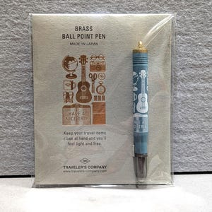 May include: A brass ballpoint pen with a blue barrel featuring travel-themed illustrations and the text "HAVE A NICE TRIP." The pen is packaged with a light grey card that includes travel-related icons and the text "BRASS BALL POINT PEN MADE IN JAPAN."