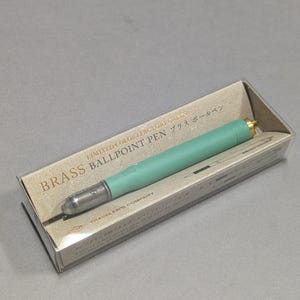 Könnte beinhalten: Ein mintgrüner Kugelschreiber aus Messing in einer transparenten Kunststoffbox. Der Stift hat eine silberne Spitze und einen goldfarbenen Akzent. Die Verpackung enthält den Text "LIMITED COLOR/FACTORY GREEN" und "BRASS BALLPOINT PEN".