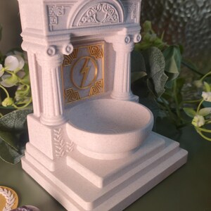 May include: A white, architectural-style decorative bowl with a gold lightning bolt emblem. The bowl is supported by columns and a tiered base, evoking classical design. The bowl is likely for decorative purposes.