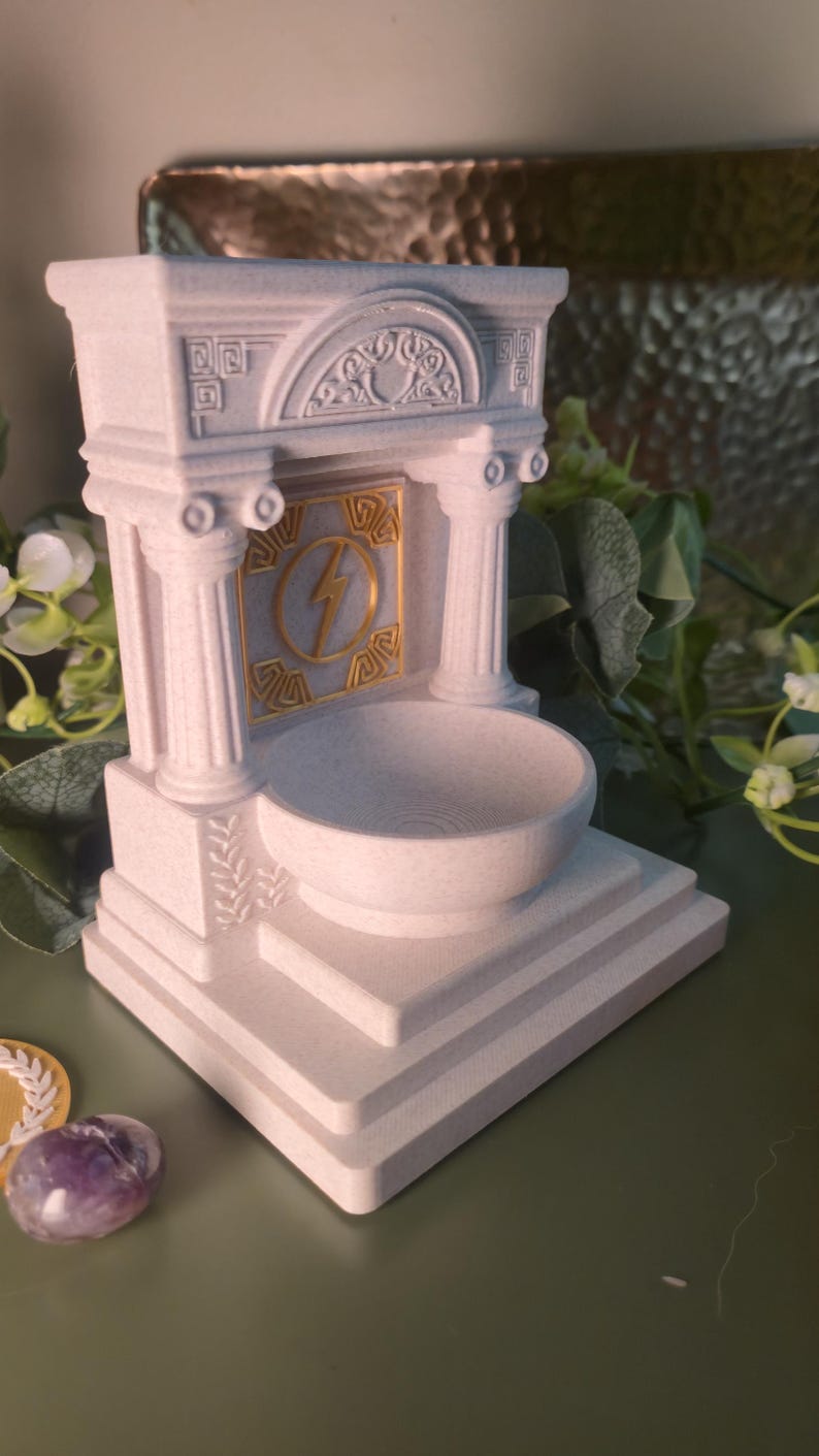 May include: A white, architectural-style decorative bowl with a gold lightning bolt emblem. The bowl is supported by columns and a tiered base, evoking classical design. The bowl is likely for decorative purposes.