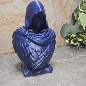 Puede incluir: Un busto azul oscuro impreso en 3D de una figura encapuchada que lleva una capa con texturas detalladas similares a plumas. El rostro de la figura está parcialmente oculto bajo la capucha y se asienta sobre una base rectangular.