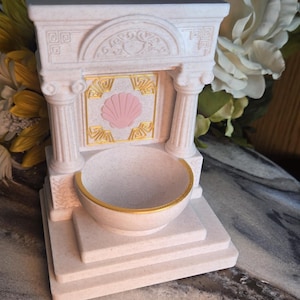 May include: A light beige, speckled, miniature fountain-shaped object featuring two columns, a central square panel with a pink seashell relief, gold-painted geometric accents, and a rounded basin with a gold-painted rim on a tiered base.