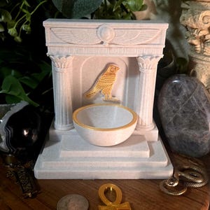 Puede incluir: Un altar decorativo blanco y dorado con un emblema de pájaro dorado, un cuenco pequeño con borde dorado y un símbolo ankh dorado. El altar tiene dos columnas y una parte superior detallada. Otros elementos incluyen una moneda de plata y una serpiente.