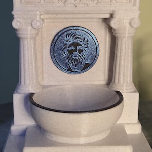 May include: A white speckled fountain featuring a central circular medallion with a blue metallic relief of a bearded face. The structure includes two fluted columns, a decorative pediment with leaf carvings, and a semi-circular basin at the base.