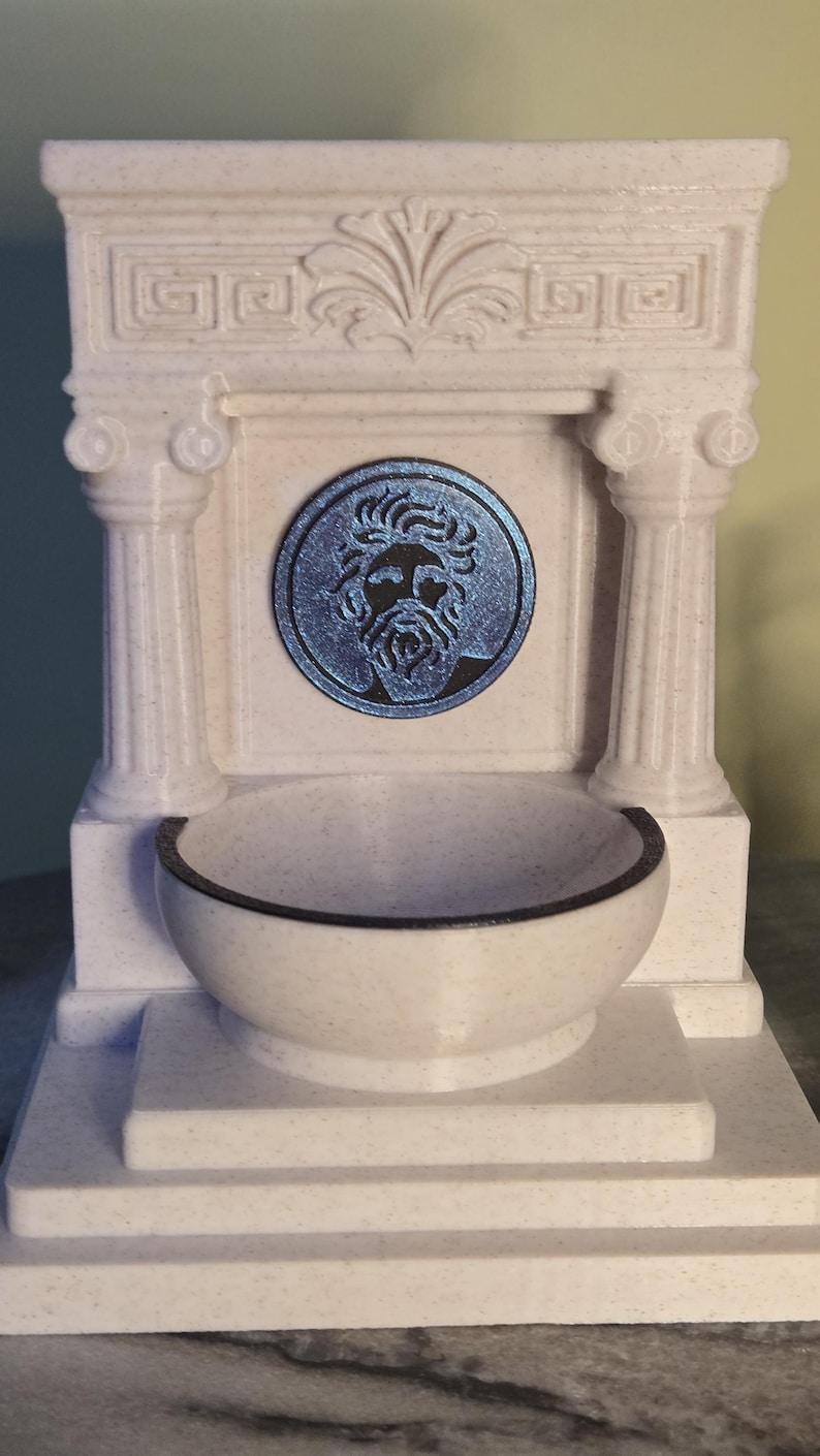 May include: A white speckled fountain featuring a central circular medallion with a blue metallic relief of a bearded face. The structure includes two fluted columns, a decorative pediment with leaf carvings, and a semi-circular basin at the base.