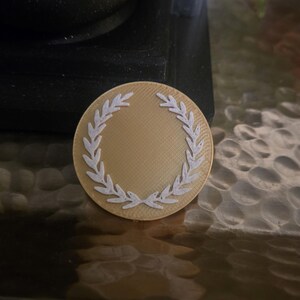 May include: A gold-colored, circular medallion with a white laurel wreath design around the edge. The medallion is set on a textured, metallic surface, with a dark background.