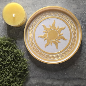 May include: A round tray featuring a gold sun emblem at the center surrounded by a gold Greek key border on a white background. The tray is positioned next to a yellow candle and a cluster of green moss, with a partial view of another tray.