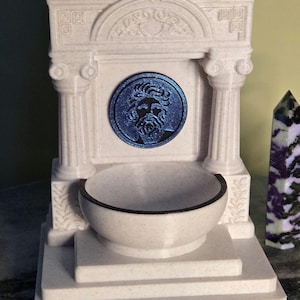 May include: A white, speckled, architectural fountain structure featuring two columns, a pediment with carved details, and a central circular medallion depicting a bearded face in metallic blue. A basin sits at the base of the structure.