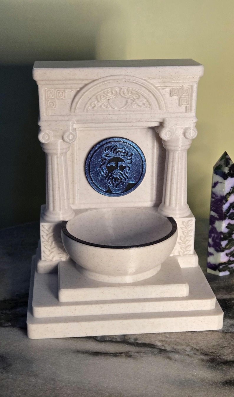 May include: A white, speckled, architectural fountain structure featuring two columns, a pediment with carved details, and a central circular medallion depicting a bearded face in metallic blue. A basin sits at the base of the structure.