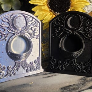 Puede incluir: Dos candelabros decorativos con la parte superior arqueada. Uno es de color púrpura claro, el otro negro. Ambos presentan un diseño de árbol de la vida con una luna creciente y una abertura circular. Los candelabros se exhiben sobre una superficie con vetas de mármol.