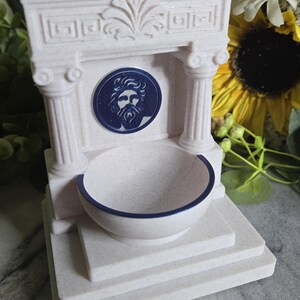 May include: A cream-colored, speckled fountain sculpture featuring a classical architectural design with two columns, a pediment, and a central circular medallion depicting a bearded face in dark blue. A semi-circular basin sits at the base.