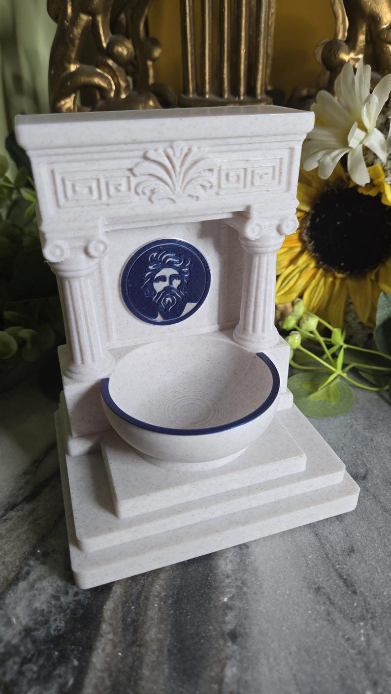 May include: A cream-colored, speckled fountain sculpture featuring a classical architectural design with two columns, a pediment, and a central circular medallion depicting a bearded face in dark blue. A semi-circular basin sits at the base.