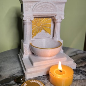 May include: A decorative, off-white structure with a sun and mountain design in yellow. It features a bowl with a gold rim, a lit orange candle, and a gold coin with a laurel wreath design. The theme is a spiritual or decorative item.