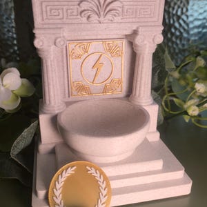 May include: A light beige miniature Greek temple with a bowl, a gold coin with a laurel wreath, and a purple stone. The temple has a gold lightning bolt symbol. The items are on a green surface.