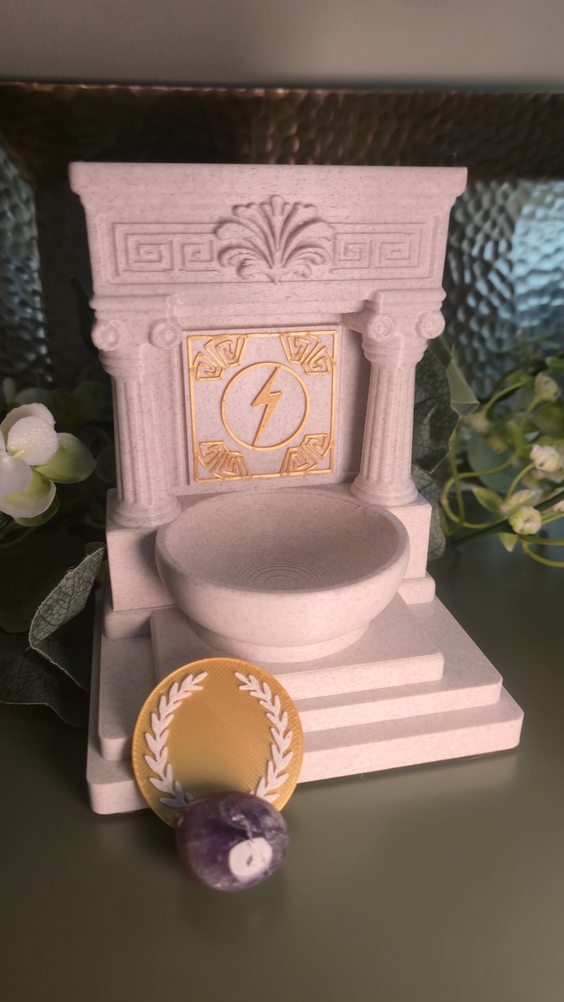 May include: A light beige miniature Greek temple with a bowl, a gold coin with a laurel wreath, and a purple stone. The temple has a gold lightning bolt symbol. The items are on a green surface.