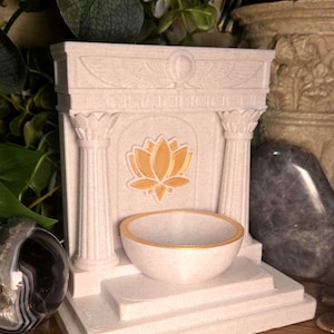Puede incluir: Un altar color crema con un diseño de flor de loto, dos columnas y un cuenco pequeño con borde dorado. El altar está sobre una superficie de madera con otros objetos decorativos.