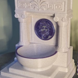 May include: A cream-colored, speckled fountain structure featuring two columns, a decorative pediment, and a central circular medallion depicting a bearded face in purple. A matching purple-rimmed basin sits at the base of the structure.