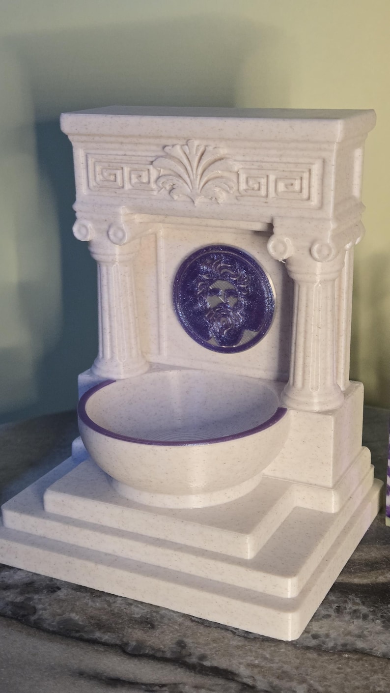 May include: A cream-colored, speckled fountain structure featuring two columns, a decorative pediment, and a central circular medallion depicting a bearded face in purple. A matching purple-rimmed basin sits at the base of the structure.