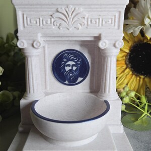 May include: A cream-colored, speckled fountain-shaped object featuring two columns, a central circular medallion with a blue relief of a bearded man's face, and a basin with a dark blue rim. The structure includes a decorative pediment top.