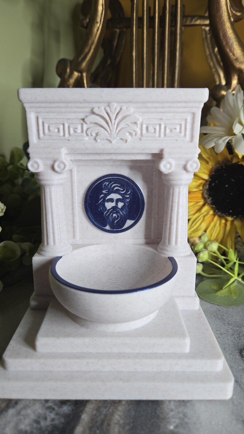 May include: A cream-colored, speckled fountain-shaped object featuring two columns, a central circular medallion with a blue relief of a bearded man's face, and a basin with a dark blue rim. The structure includes a decorative pediment top.