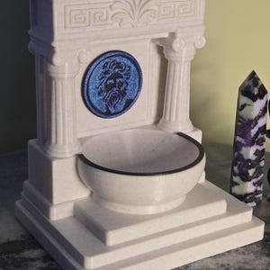 May include: A cream-colored, speckled fountain structure featuring two columns, a central circular blue medallion with a relief of a bearded face, and a basin. The architectural design includes a stepped base and a decorative pediment top.