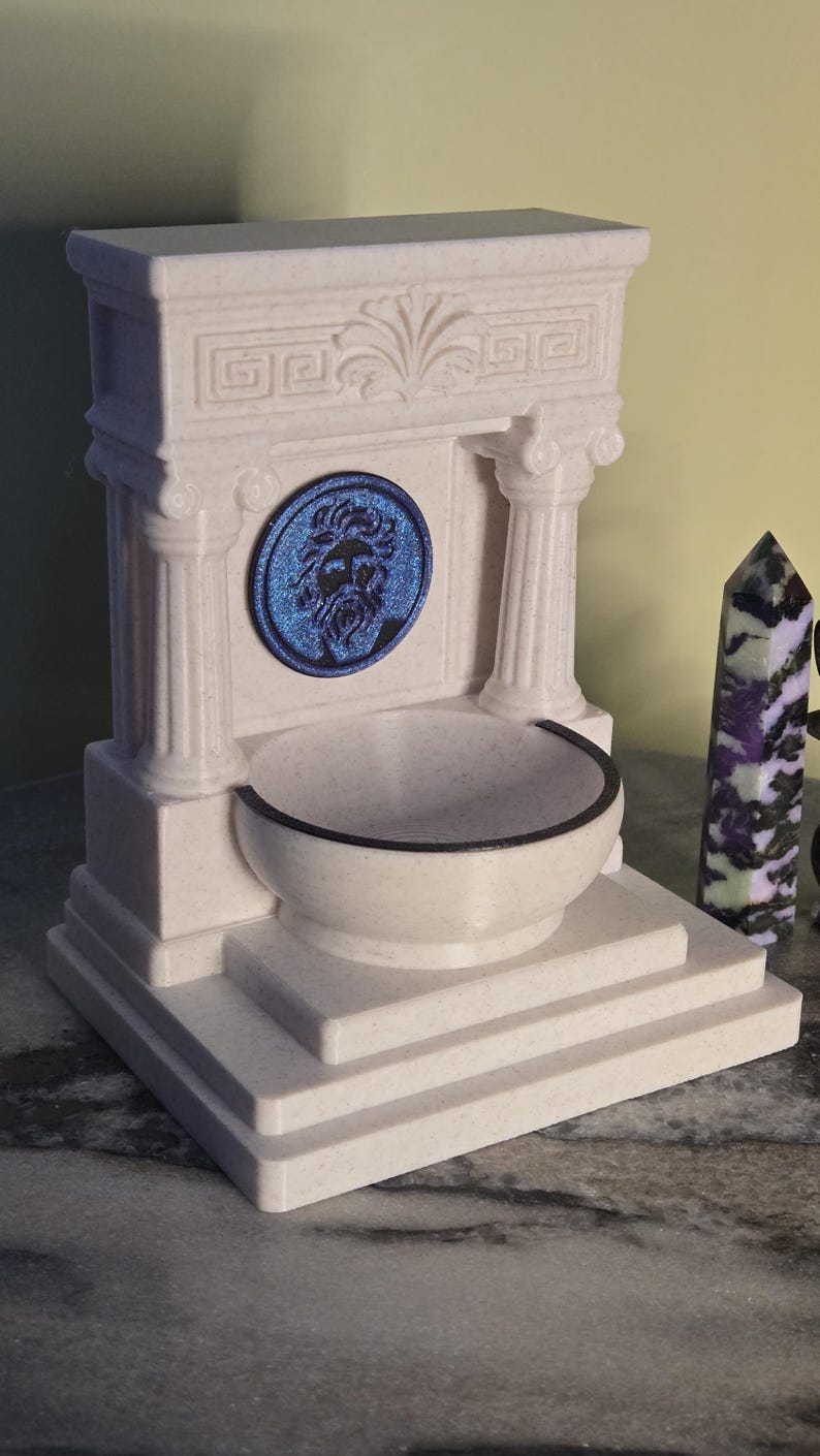 May include: A cream-colored, speckled fountain structure featuring two columns, a central circular blue medallion with a relief of a bearded face, and a basin. The architectural design includes a stepped base and a decorative pediment top.