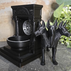 Puede incluir: Una figura de perro de tres cabezas de color negro junto a una estructura arquitectónica negra que presenta un cuenco circular y un emblema circular plateado con un patrón de laberinto en la pared trasera de la base.