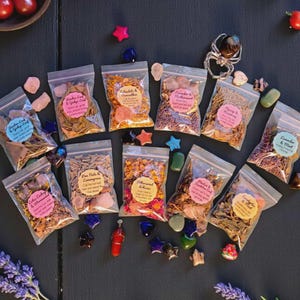 May include: An overhead shot of several small, clear plastic bags filled with dried herbs and flowers, each labeled with a round sticker. Various colorful crystals and star-shaped decorations are scattered around the bags on a dark surface.