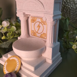 May include: A white, architectural-style decorative piece with a bowl, columns, and a lightning bolt emblem. A gold-colored laurel wreath and a purple stone are in front of the structure. The piece has a classical design.